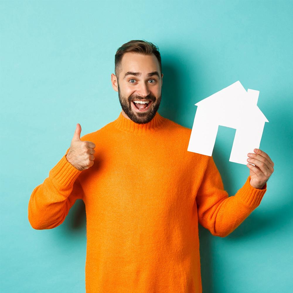 Photo d'un homme heureux en pull orange qui tiens dans sa main une maison en carton blanche pour le slider de présentation d'agence immobilière